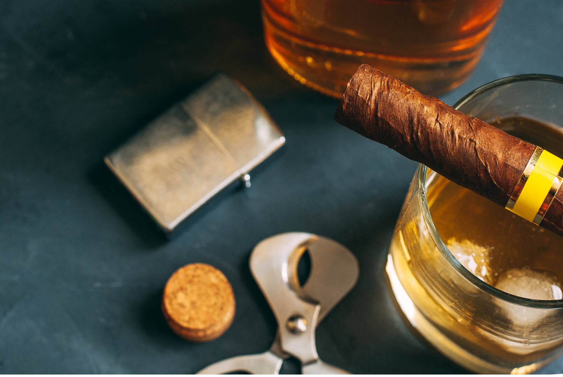 An image of quality cigars and cognac on dark dining accessories, cigar scissors and torch lighter.