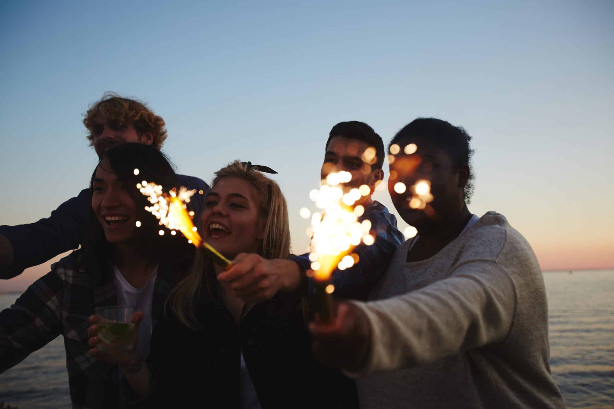 Male and female friends with sparklers lighten using disposable lighters in hands gathered together outdoors and celebrating momentous event, picturesque seascape on background